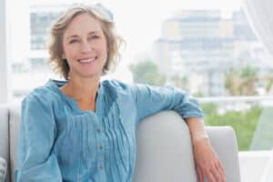 50-year-old woman smiles while sitting on the couch, mitigating her menopause symptoms.