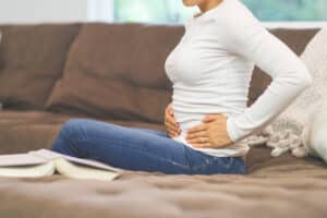 Woman sitting on the couch with her hands on her lower stomach, suffering from symptoms of pelvic prolapse.